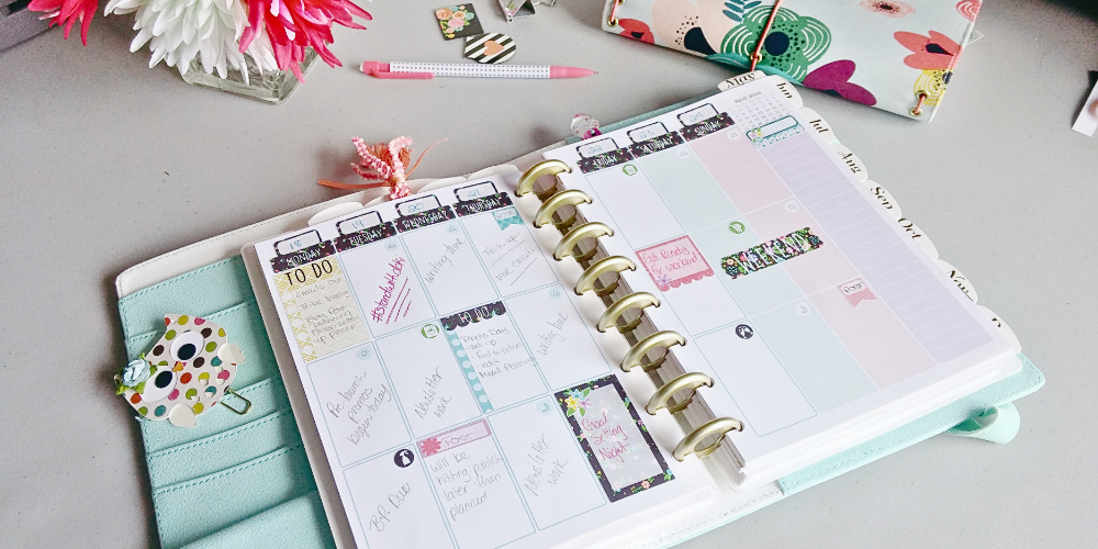 Here is a top down picture of a desk. The main part of the image shows a teal and white discbound planner lying open to a weekly planner spread. The week is planned out using stickers and colour pens. The left hand side of the planner shows a column of pockets with a paper, rainbow polka dotted owl paper clip. Above the planner from left to right are: a square cut glass vase with white and pink flowers in in, a white polka dot pencil with pink ends, some magnetic bookmarks, and a closed, cloth travellers notes book. The fabric for that book is colourful, doodle like flowers on a pale blue background. The elastic strings are pink.