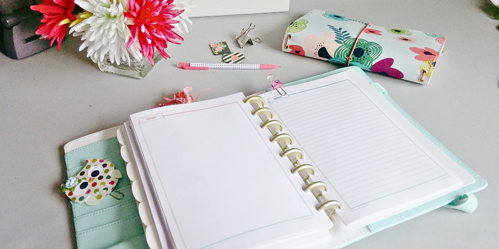 Here is a top down picture of a desk. The main part of the image shows a teal and white discbound planner lying open to a to show a dot grid page labeled ideas on the left and a lined page titled notes on the right. The left hand side of the planner shows a column of pockets with a paper, rainbow polka dotted owl paper clip. Above the planner from left to right are: a square cut glass vase with white and pink flowers in in, a white polka dot pencil with pink ends, some magnetic bookmarks, and a closed, cloth travellers notes book. The fabric for that book is colourful, doodle like flowers on a pale blue background. The elastic strings are pink.