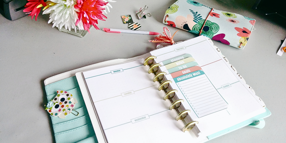 Here is a top down picture of a desk. The main part of the image shows a teal and white discbound planner lying open to a weekly planner spread. The week is a Monday start, horizontal layout on two pages. On the right hand side, in a series of lists paged attached to the discs in the centre. Top to bottom they read, shopping, notes, to-do, goals, and absolutely must. The left hand side of the planner shows a column of pockets with a paper, rainbow polka dotted owl paper clip. Above the planner from left to right are: a square cut glass vase with white and pink flowers in in, a white polka dot pencil with pink ends, some magnetic bookmarks, and a closed, cloth travellers notes book. The fabric for that book is colourful, doodle like flowers on a pale blue background. The elastic strings are pink.